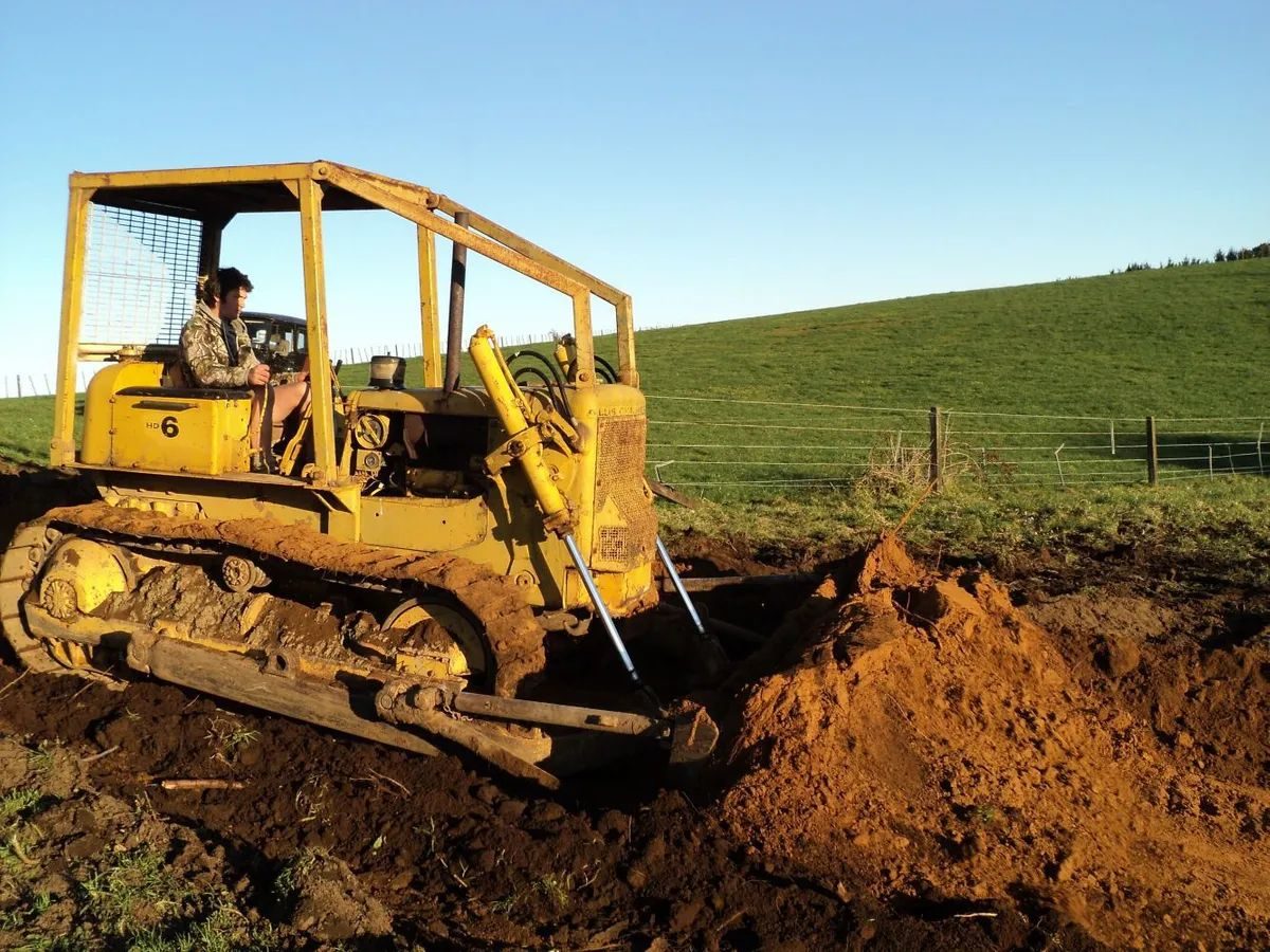 Operator on an older yellow bulldozer with a roll cage, pushing up red-brown soil at the foot of a grassy slope