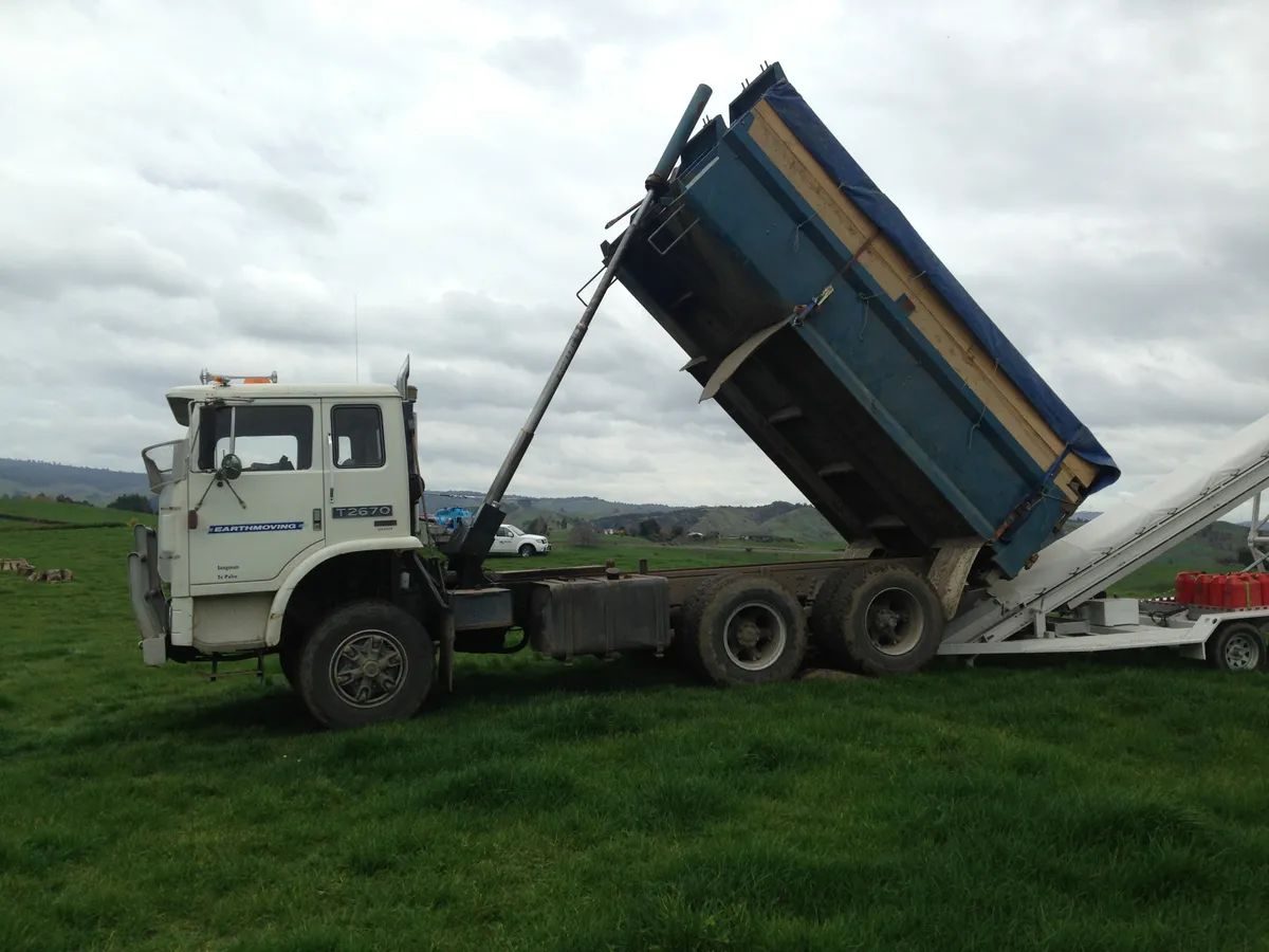 White truck with its blue tipping bin raised, standing in a grass paddock under a cloudy sky