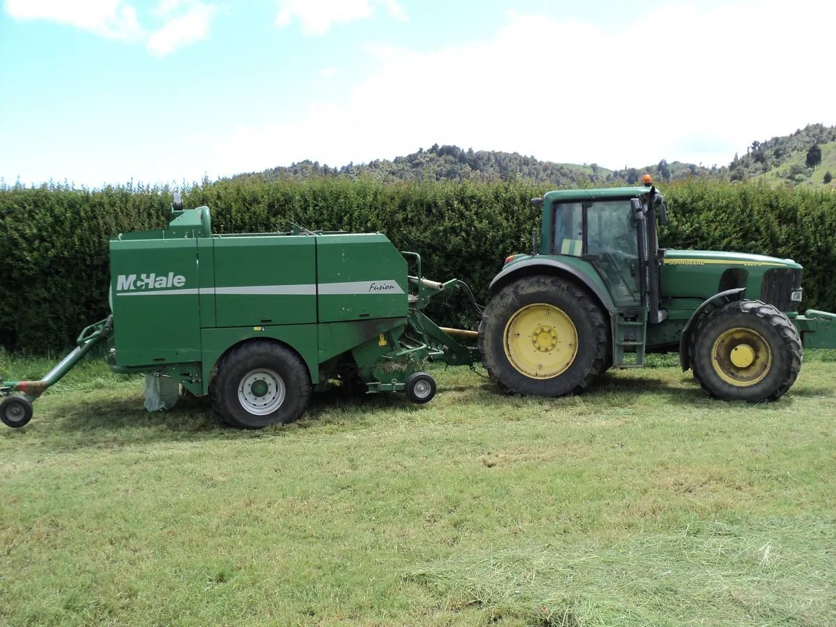 Green McHale baler hitched to a John Deere tractor, parked on short grass in front of a tall hedge